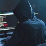 Person in hoodie using computer with code on screen.
