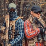 Two armed individuals in tactical gear in forest.