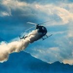 Helicopter releasing smoke over mountainous landscape with clouds