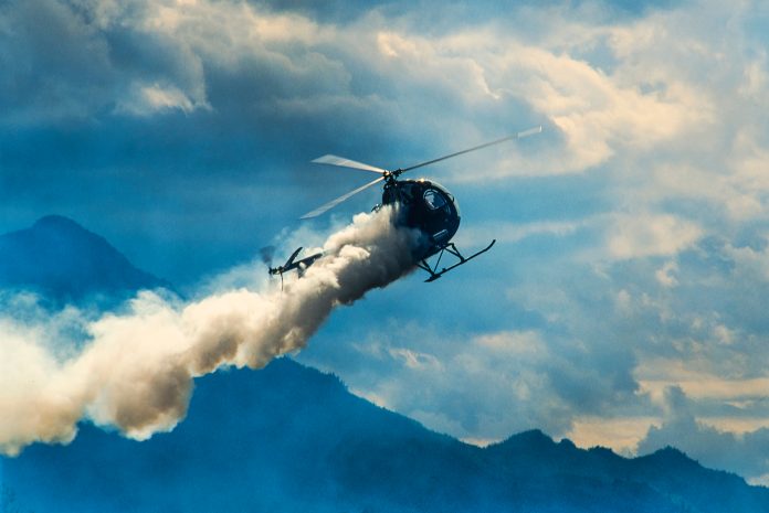 smoking helicopter in flight_1429549637.jpg featured image Helicopter releasing smoke over mountainous landscape with clouds