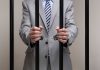 Businessman in a suit holding prison bars with handcuffs on his wrists