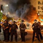 DEADLY PROTESTS — President Won’t Step Down! Police officers in riot gear near burning car.