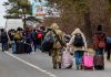 Asylum Scandal UNCOVERED – Government Wasted BILLIONS! Group of people carrying luggage walking along a road with a soldier