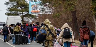 Group of people carrying luggage walking along a road with a soldier