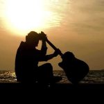 Silhouette of a guitarist sitting by the ocean during sunset