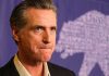 Newsom’s Top Advisor ARRESTED — $225K THEFT Man in suit, solemn expression, purple background with bear drawing.