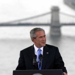 A man in a suit giving a speech at a podium with a bridge in the background