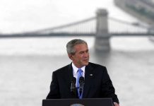 Bush BREAKS Silence – Offers Glowing Tribute A man in a suit giving a speech at a podium with a bridge in the background