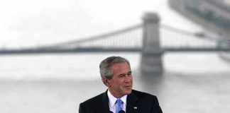 A man in a suit giving a speech at a podium with a bridge in the background