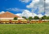 A row of yellow school buses parked in front of a school building under a cloudy sky