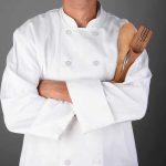 A chef in a white uniform holding wooden cooking utensils with crossed arms