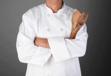 A chef in a white uniform holding wooden cooking utensils with crossed arms