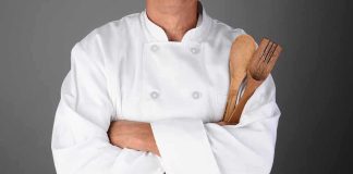 A chef in a white uniform holding wooden cooking utensils with crossed arms