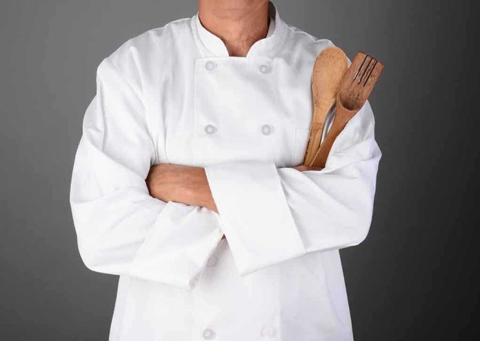 A chef in a white uniform holding wooden cooking utensils with crossed arms