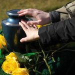 Hands touching an urn surrounded by yellow roses.