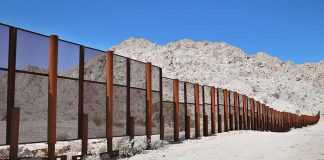 A long border wall stretches across a desert landscape with mountains in the background