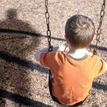 A young boy sitting on a swing at a playground, with his shadow cast on the ground