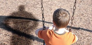 A young boy sitting on a swing at a playground, with his shadow cast on the ground