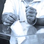 A handcuffed individual placing a ballot into a transparent voting box