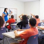 Teacher in a blue dress instructing students in a classroom with hands raised