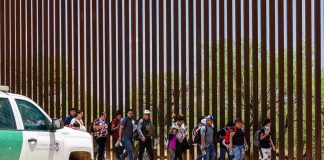 Border Wall Deadline Sparks 2027 Showdown People walking beside tall fence and border patrol vehicle.