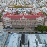 Aerial view of a large historic school building with red roof in a dense city neighborhood