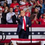 man in suit points from a stage at a political rally
