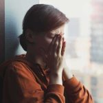 person in orange hoodie covering face by window