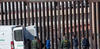 Border agents escort a group near a tall fence and a patrol van