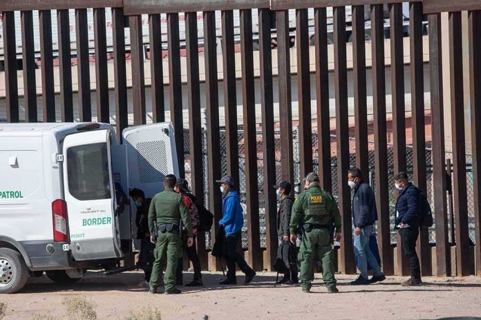 Border agents escort a group near a tall fence and a patrol van