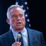 Watch RFK Jr TORCH Democrats’ Covid Response On Stage Man holding microphone, speaking with American flag behind.
