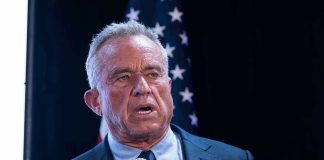 Watch RFK Jr TORCH Democrats’ Covid Response On Stage Man holding microphone, speaking with American flag behind.