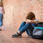 America’s First Measles Outbreak of 2026 Turns DEADLY boy sitting against brick wall with head on arms and backpack
