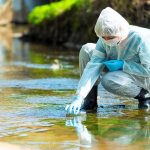 Scientist in protective suit collecting water sample from stream