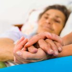 Hospice Scam Explosion — Taxpayers Bleed Caregiver holding hands with an elderly patient in bed