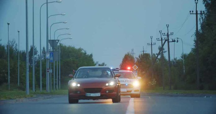 red car driving on road with police car behind using flashing lights