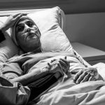 Six-Month Rule Ignites Assisted-Dying Backlash Patient in hospital bed with nasal oxygen held by visitor