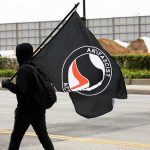 Hooded person carrying a large black flag on a city street