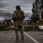Soldier in camouflage stands at roadblock with smoke in the distance
