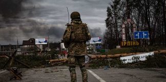 Soldier in camouflage stands at roadblock with smoke in the distance