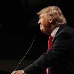 Donald Trump speaking at a podium in profile against a dark background