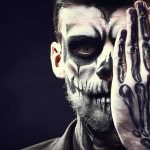 Person with skull face paint covering one eye with a skeleton-painted hand