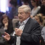 George Soros speaks with a microphone; Viola Odebrecht is seated in the audience