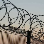Razor wire coils atop a metal security fence at dusk